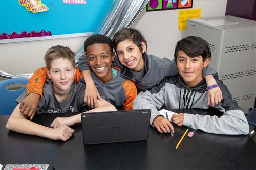 Four middle school students sitting at a table, in front of a laptop, smiling 